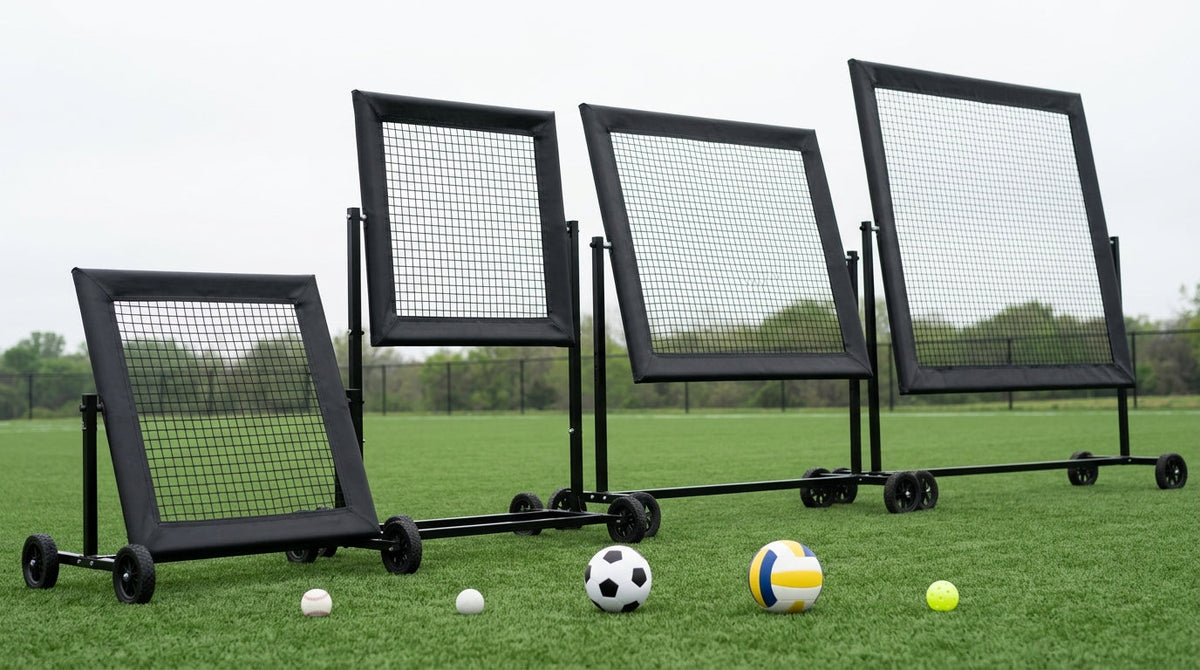 Four black All Ball Pro rebounders of increasing size lined up on turf with their screens set at different angles, and a baseball, lacrosse ball, soccer ball, volleyball and pickleball resting on the ground in front.